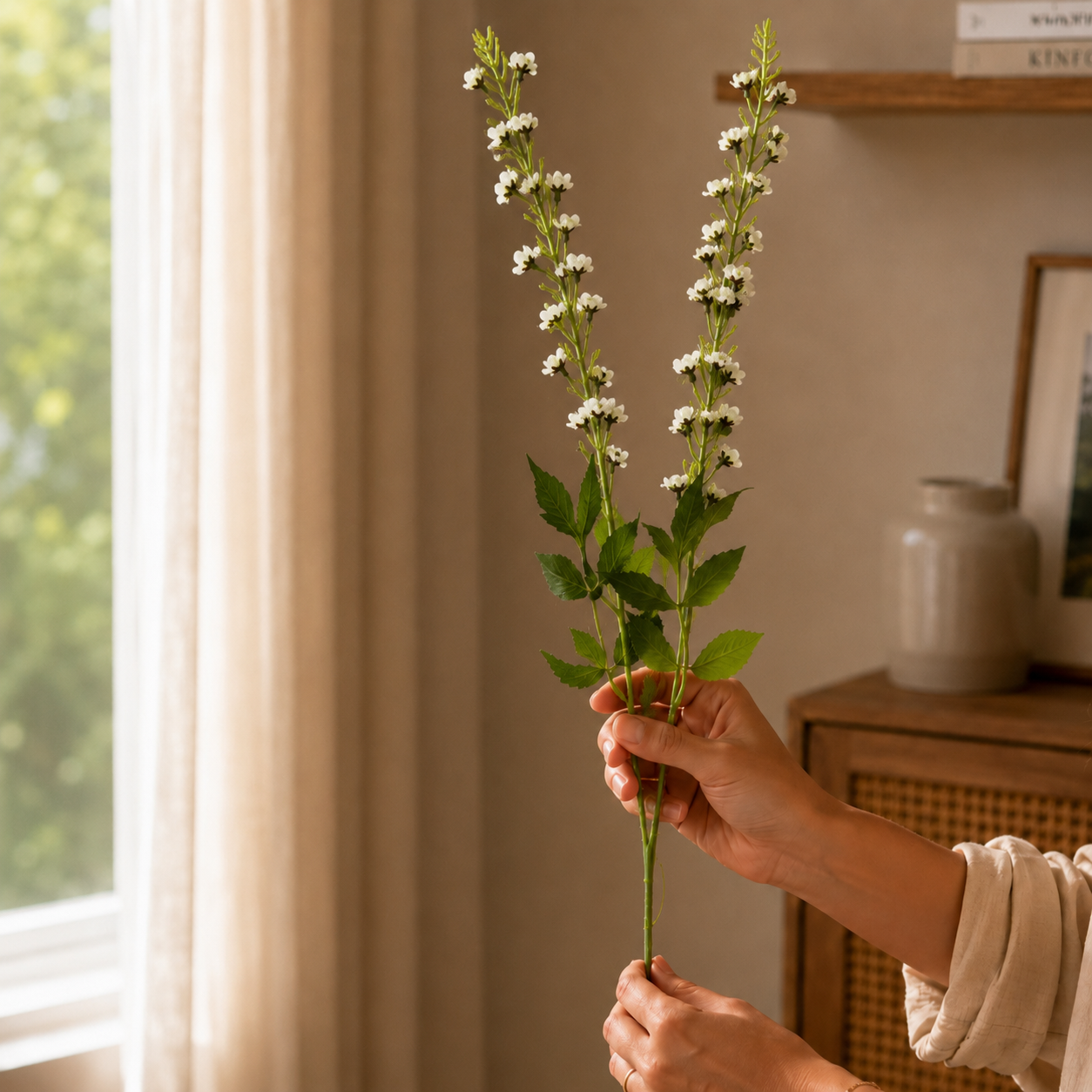 Hand holding white artificial flower stems in a warm, naturally lit living room, showcasing realistic floral details for elegant home decor styling.