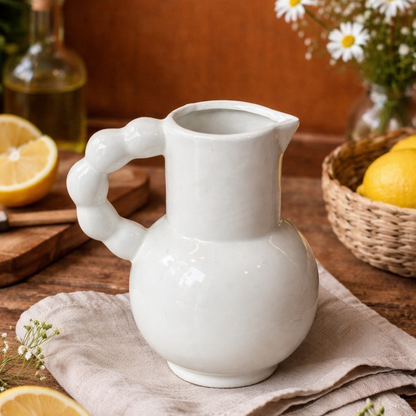 White ceramic pitcher vase with a sculpted loop handle placed on a wooden table with lemons and flowers in the background, styled in a warm kitchen setting.