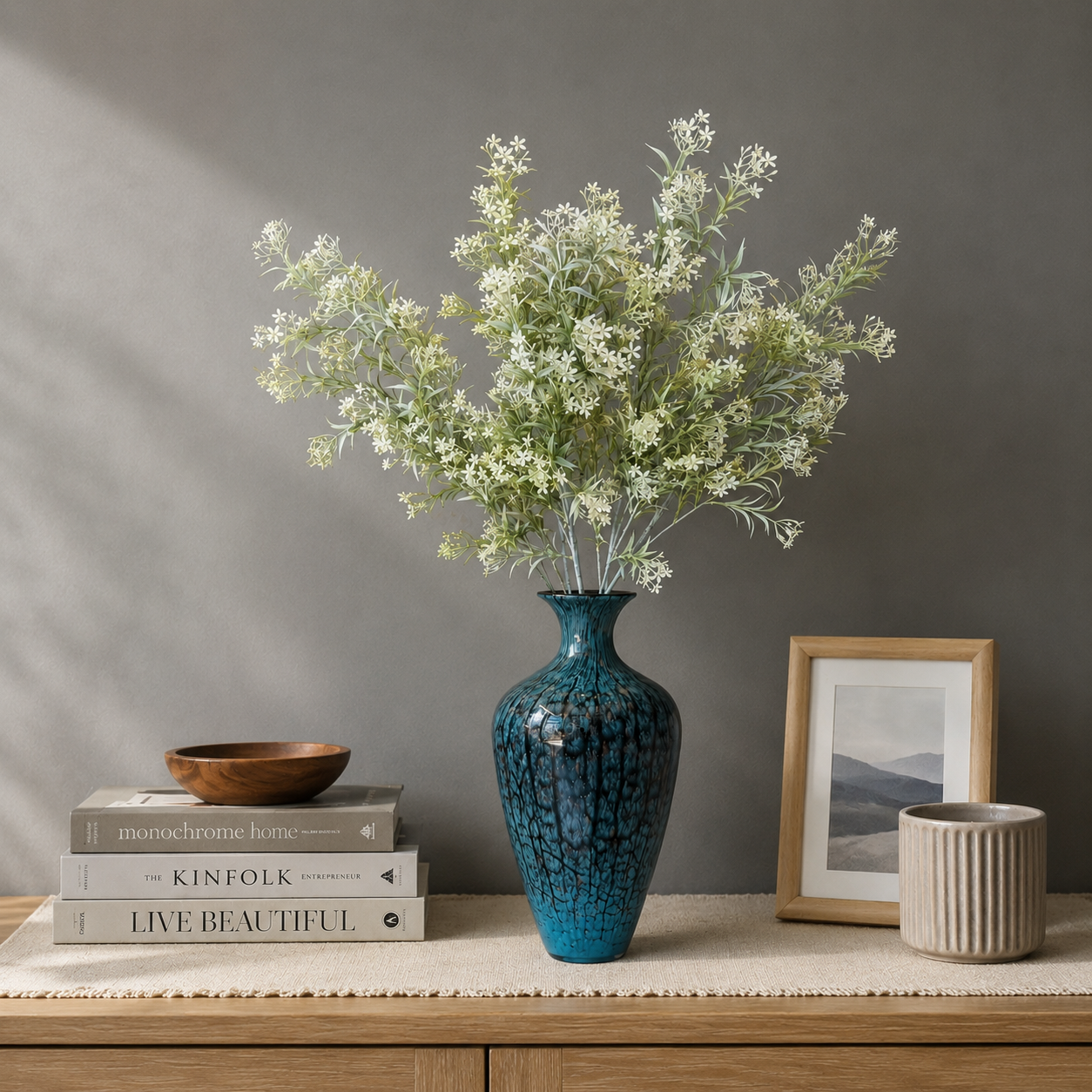 Artificial white floral arrangement displayed in a textured blue ceramic vase on a wooden table against a grey wall, styled with books and decorative accents in a modern home interior.