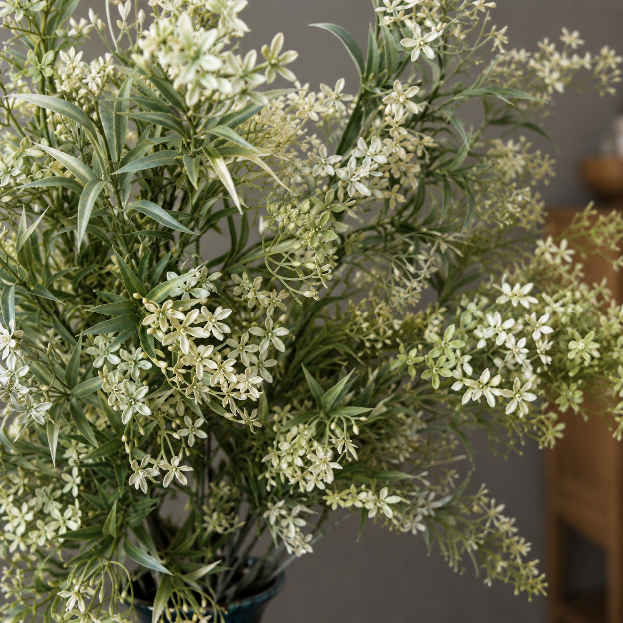Close-up lifestyle view of a white star floral bouquet with soft green foliage arranged in a dark blue vase, styled on a wooden table against a grey wall in a modern home décor setting.