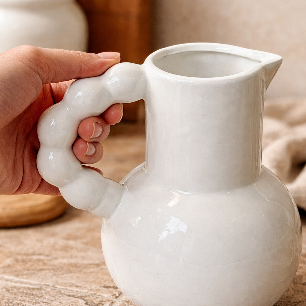 A detailed close-up shot of a hand holding the ergonomic, rounded bubble handle of a white stoneware pitcher.