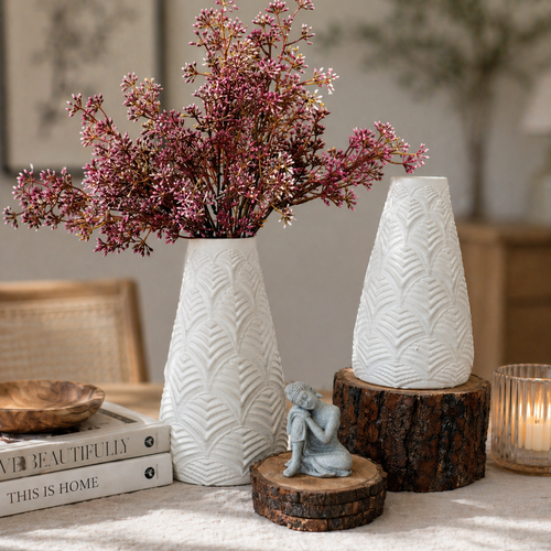 Close-up lifestyle image of textured white ceramic vases with pink floral stems styled on a wooden tabletop with rustic accents and soft natural lighting.