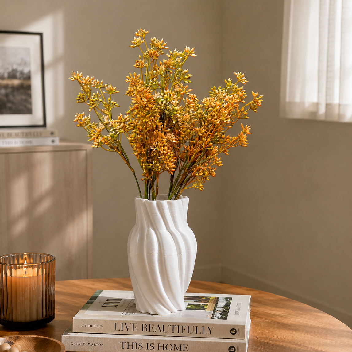 White wave-textured ceramic vase with orange floral stems displayed on stacked coffee table books in a warm minimalist living room setting with soft natural light and decorative candle accents.