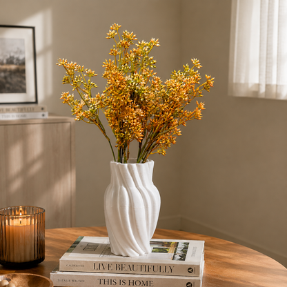 White wave-textured ceramic vase with orange floral stems displayed on stacked coffee table books in a warm minimalist living room setting with soft natural light and decorative candle accents.