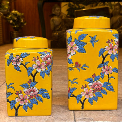 A set of two bright yellow rectangular ceramic ginger jars with hand-painted pink cherry blossoms and blue leaves.