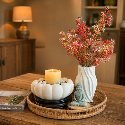 Elegant tabletop decor setup featuring a white sculpted vase with pink floral stems, a lotus-shaped candle holder with pebbles, and a Buddha figurine arranged on a woven tray in a warm modern living space.