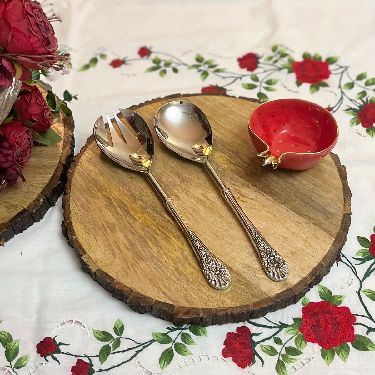 Ornate silver salad serving set resting on a round rustic wooden board, styled with a red pomegranate-shaped bowl and a rose-patterned tablecloth.