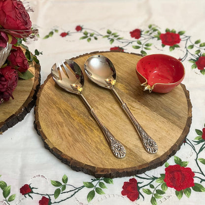 Ornate silver salad serving set resting on a round rustic wooden board, styled with a red pomegranate-shaped bowl and a rose-patterned tablecloth.
